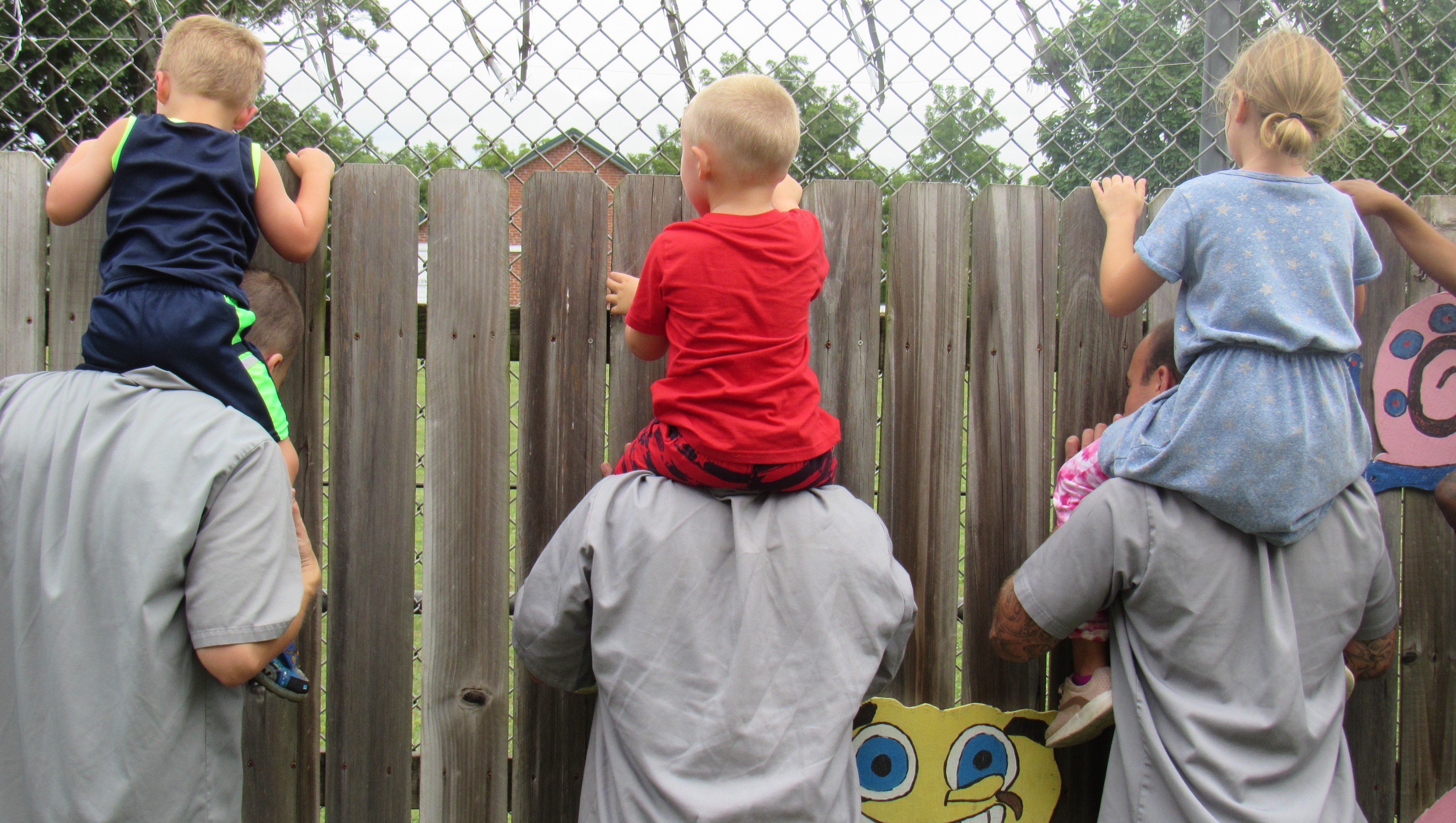 Kids sit on their dads' shoulders in an outside play area on correctional center grounds.