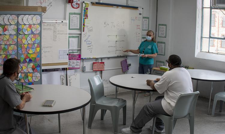 Teaching in a mask at the front of a classroom, with two inmate students at desks