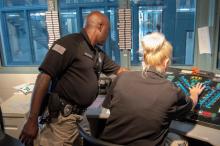 A man and a woman in corrections officer uniforms work at a control panel in a prison housing unit.